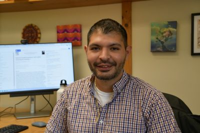 Man at desk faces camera with computer screen in the background.