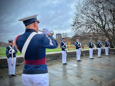 A cadet bugler plays Taps during a wreath-laying ceremony at the Pylons while cadets in dress uniform stand at attention.