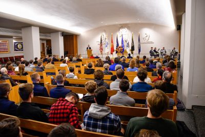  The pews of the Chapel are filled with guests and a speaker, cadet brass ensemble, and flags are on stage during the remembrance ceremony.